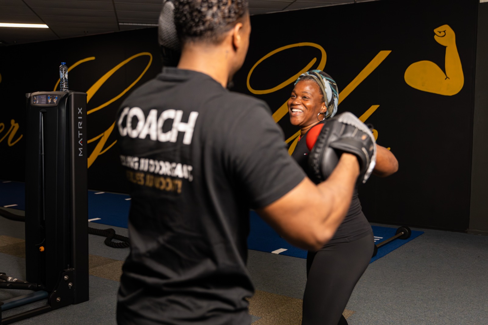 A woman smiling while boxing with a coach holding mitts in a gym with black and yellow wall art.