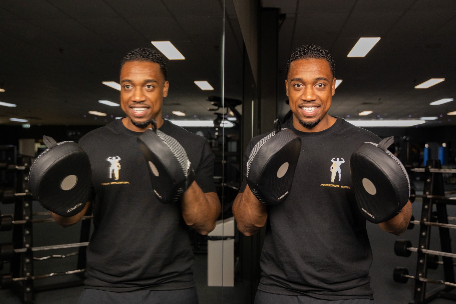 Smiling personal trainer in black shirt holding boxing focus mitts in a gym with his reflection visible in a mirror.