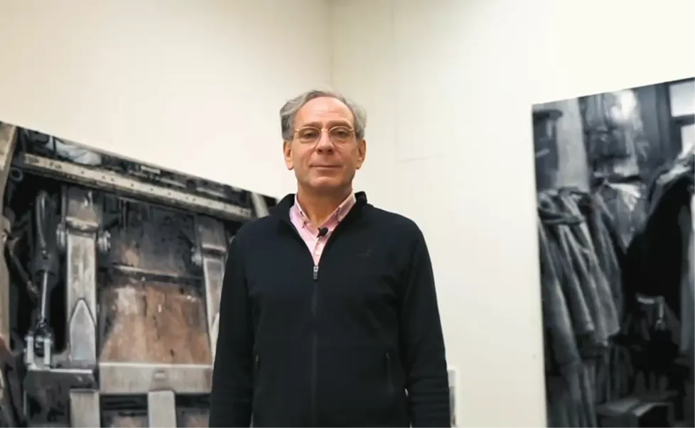 Man with glasses wearing a black zip-up sweater standing in front of large, industrial-style paintings in a gallery.