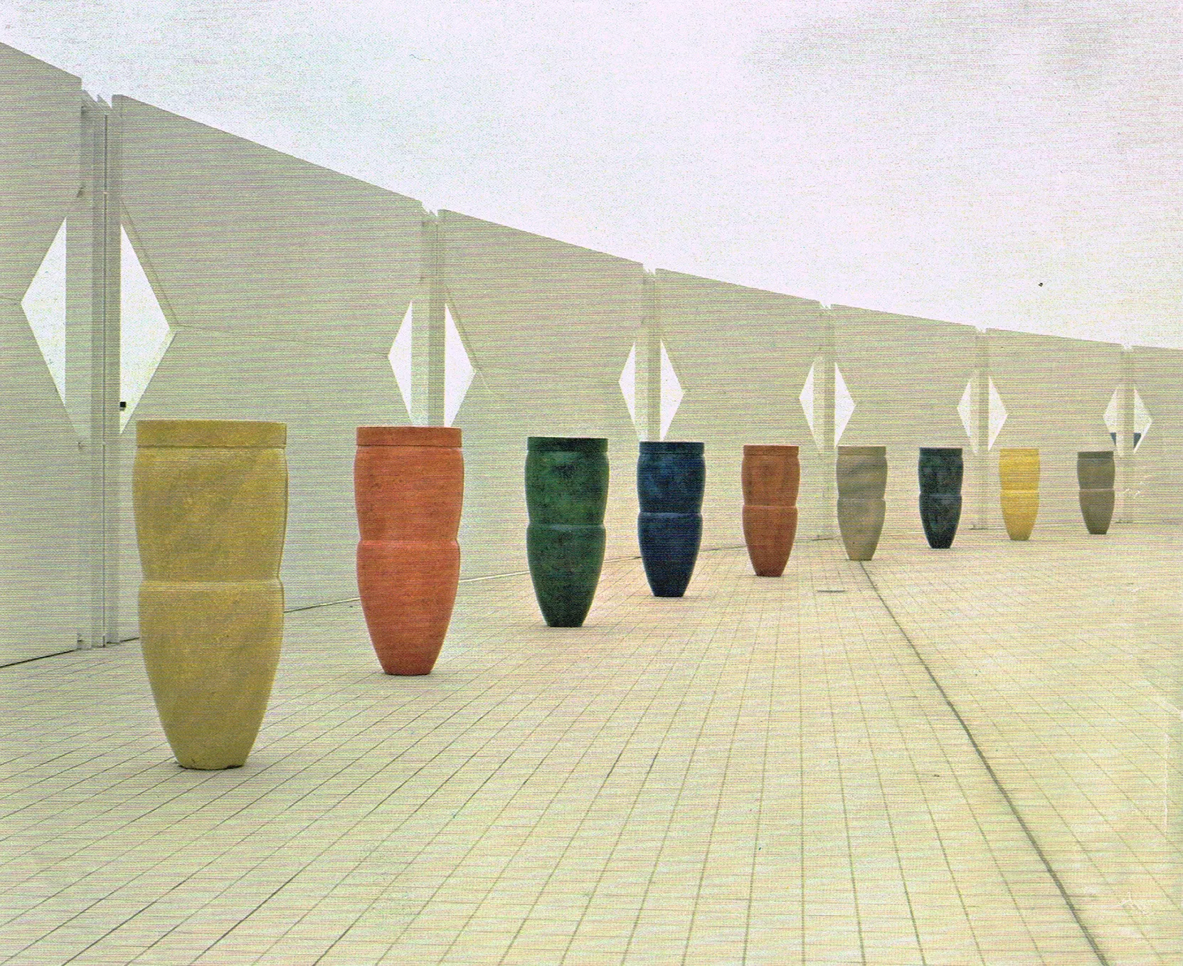 Row of tall, colorful clay vases arranged on a tiled floor against a white geometric wall.