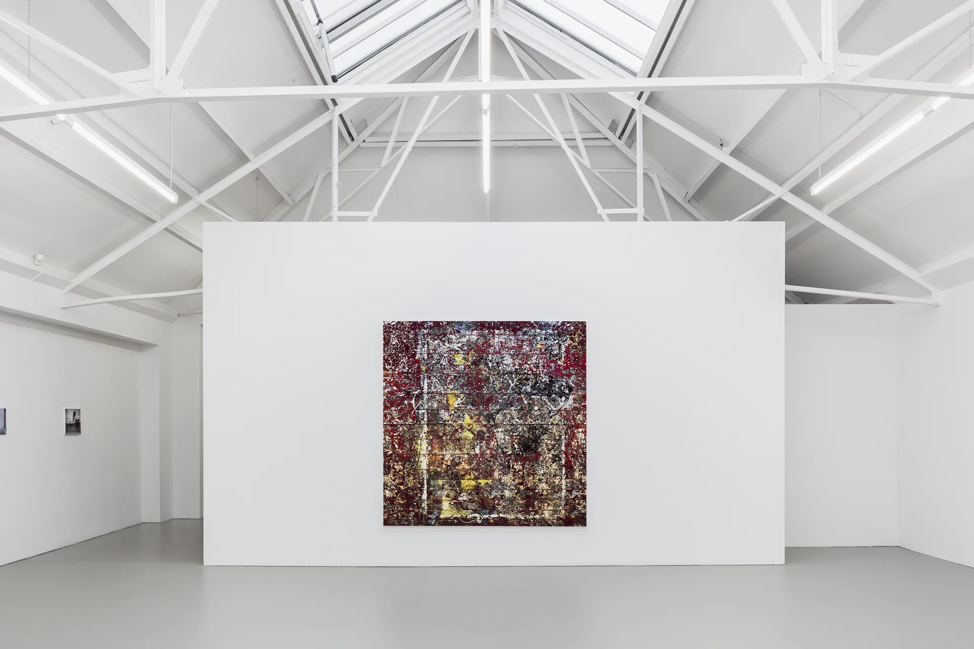 Abstract expressionist painting with splattered red, yellow, white, and black paint on a square canvas displayed on a white gallery wall.