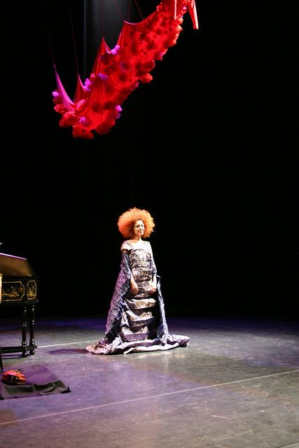 Performer with an afro hairstyle wearing a long patterned gown standing on a stage under a large red fabric installation suspended above.