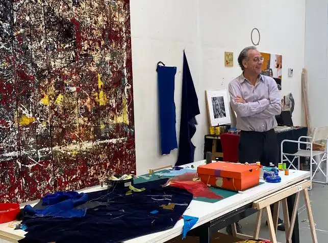 Artist in a studio standing near a table with fabric and art supplies, with a large abstract painting on the wall.