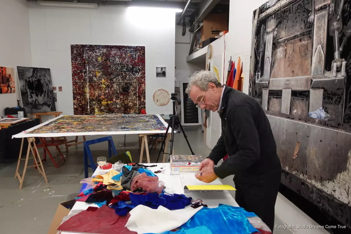 Artist working on a colorful collage at a cluttered table in a studio surrounded by abstract paintings.