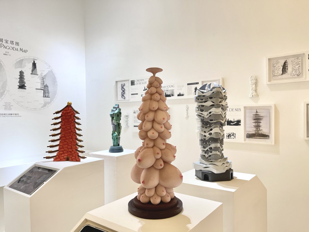 Art gallery display featuring four modern sculptures on white pedestals, including a central tower-like sculpture made of multiple realistic breast forms.