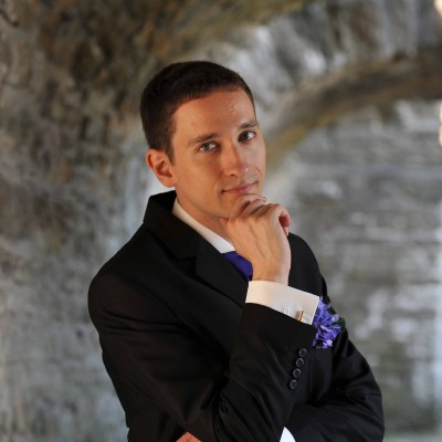 Young man in a black suit with a purple tie and boutonniere, posing thoughtfully under a stone archway.
