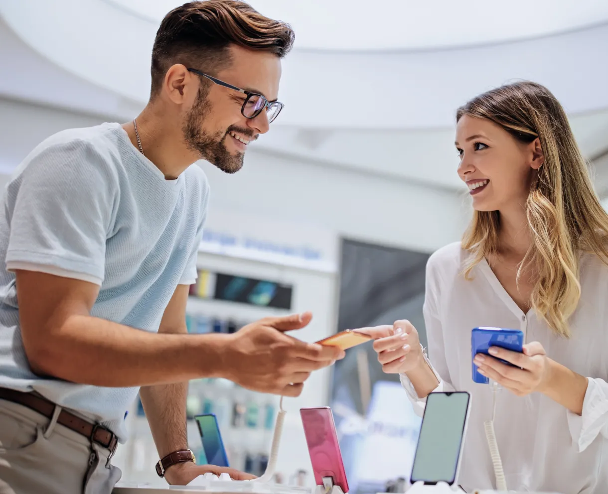 A man and woman smiling and interacting while holding smartphones in a modern electronics store.