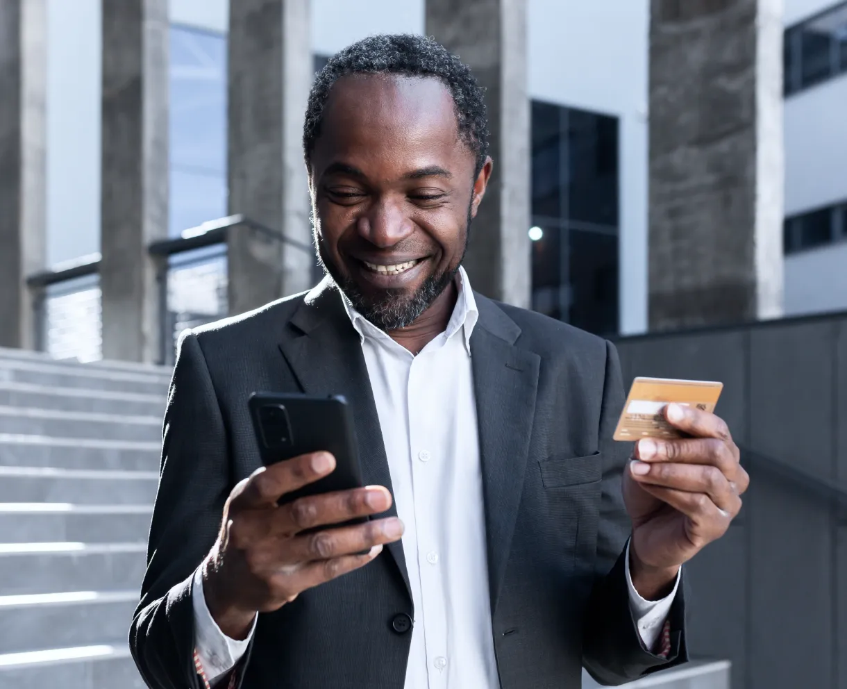 Smiling man in suit holding a credit card and looking at his smartphone outdoors.
