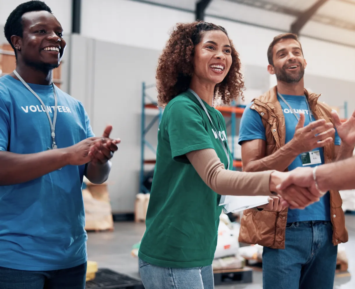 Three volunteers in a warehouse, two men and one woman, smiling and shaking hands.