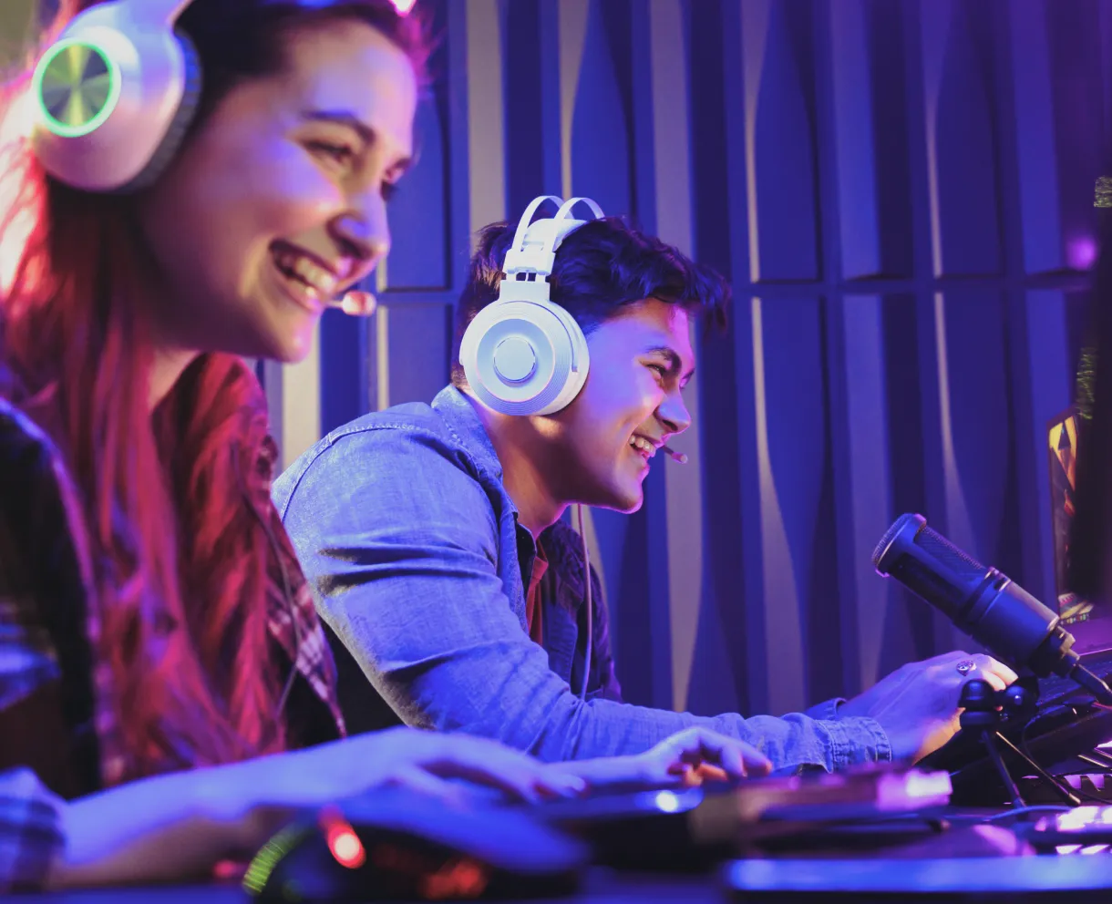Two young people wearing headsets and smiling while gaming with a microphone and computer in a colorful room.
