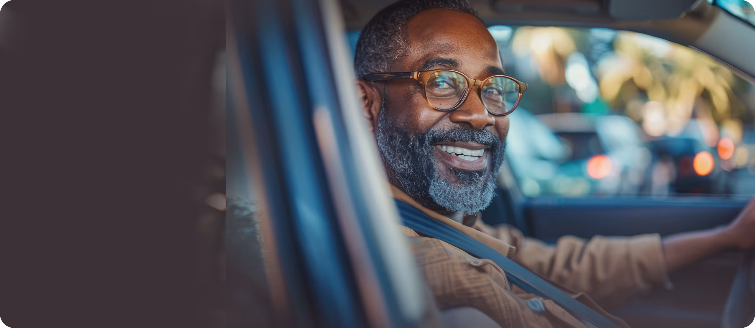 Smiling bearded man wearing glasses and a seatbelt, sitting in the driver's seat of a car.