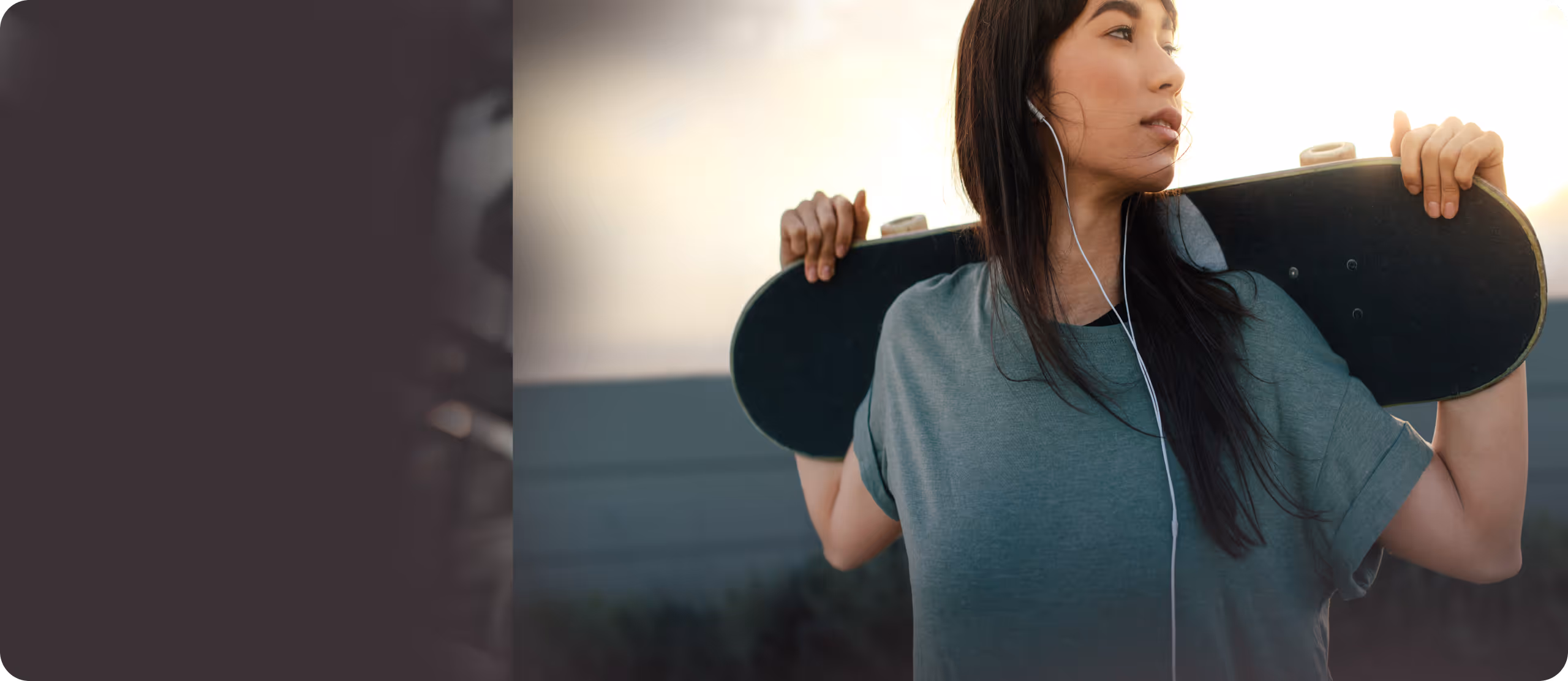 Young woman holding a skateboard behind her shoulders while listening to music with earphones outdoors at sunset.