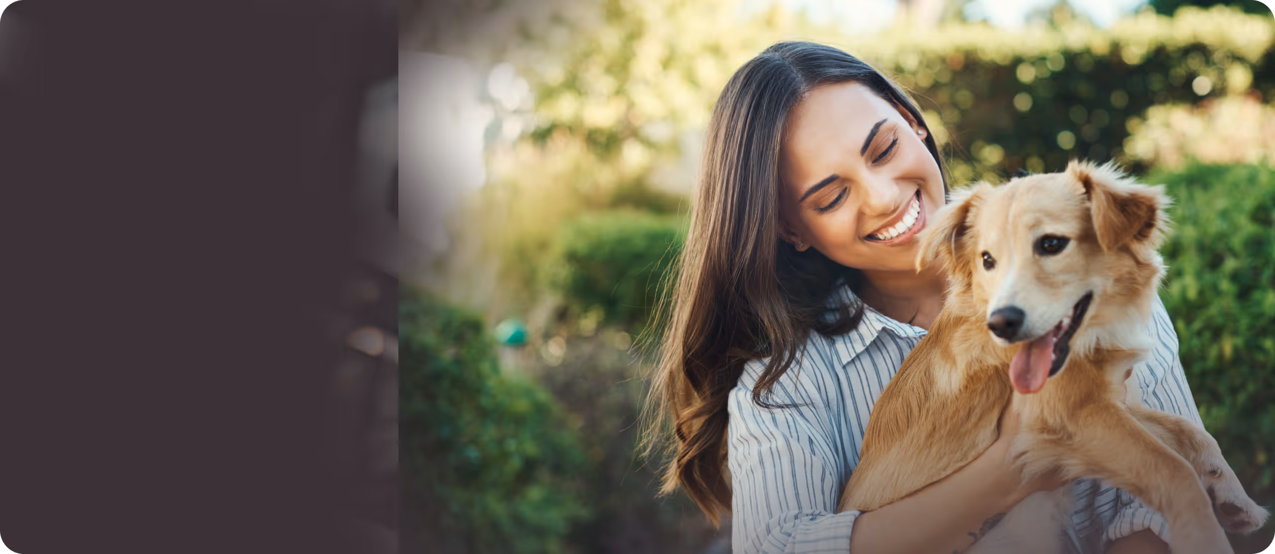 Smiling woman with long hair holding a happy light brown dog outdoors with green bushes in the background.