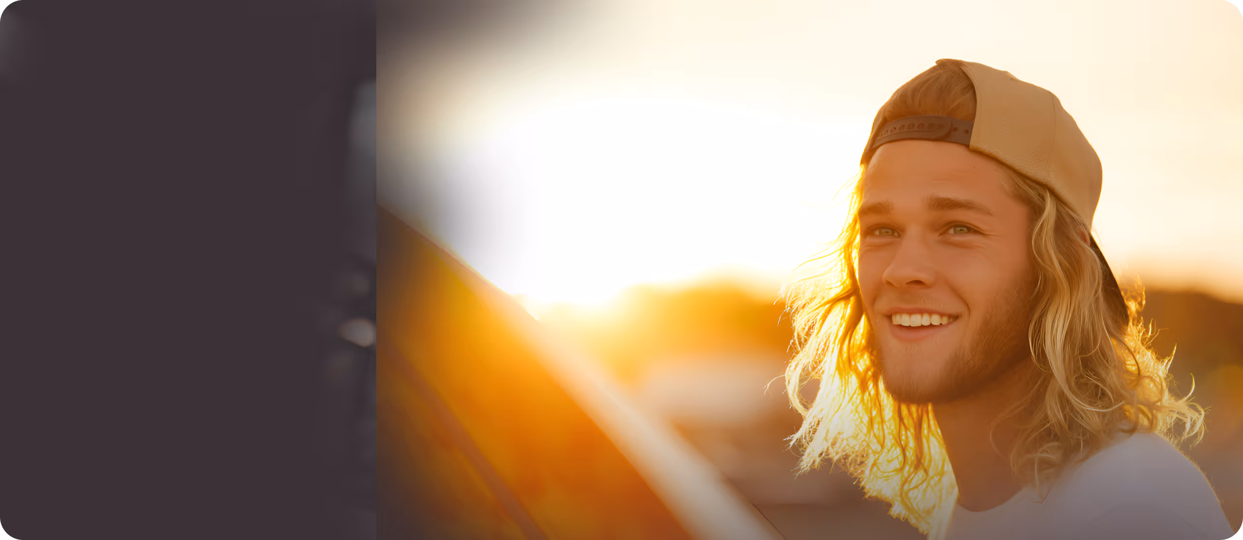 Young man with long blond hair wearing a backwards cap smiling with a sunset background.