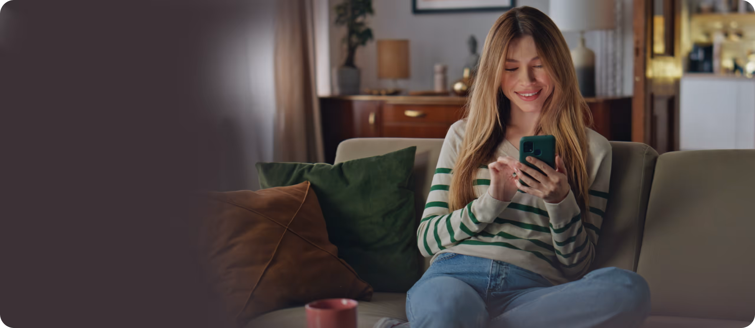 Smiling young woman sitting cross-legged on a couch while using a smartphone.