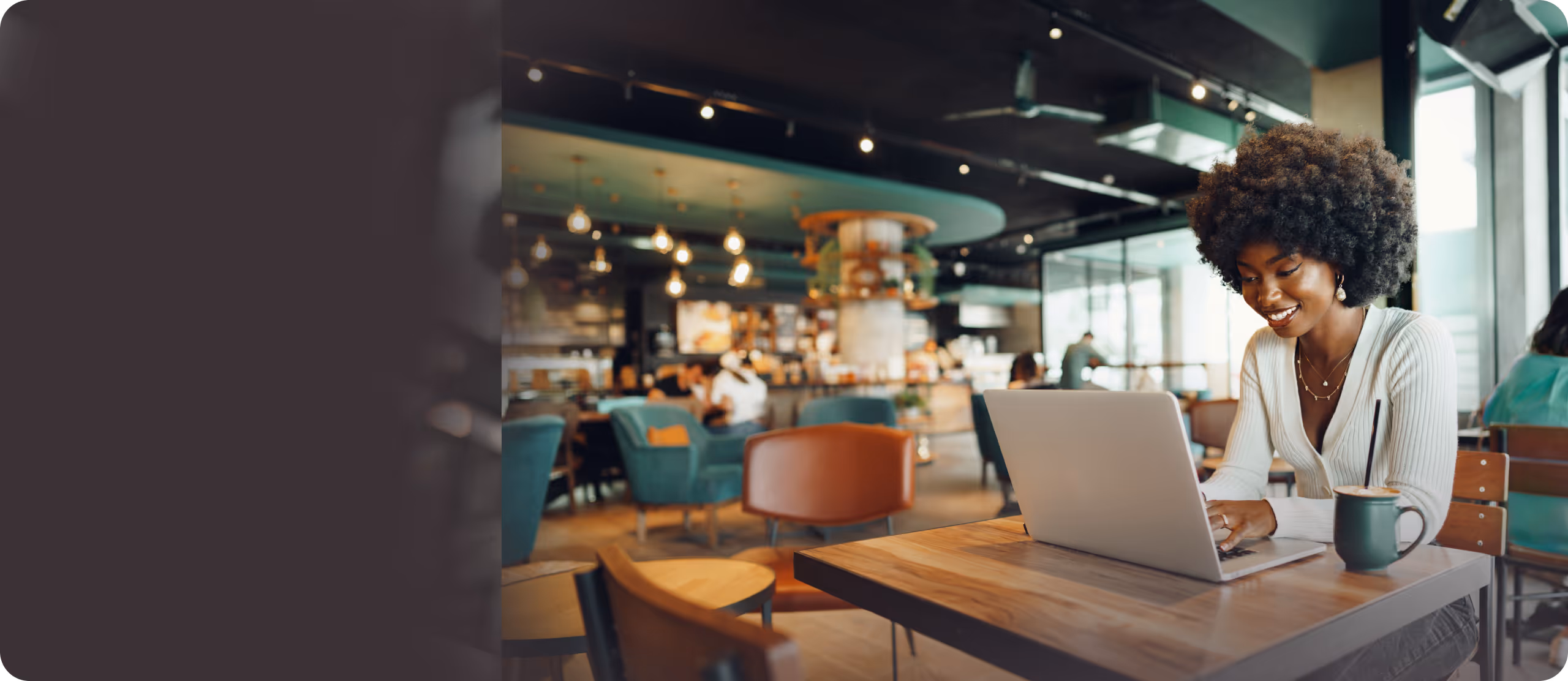 Smiling woman with curly hair working on a laptop at a wooden table in a modern café with a coffee cup beside her.