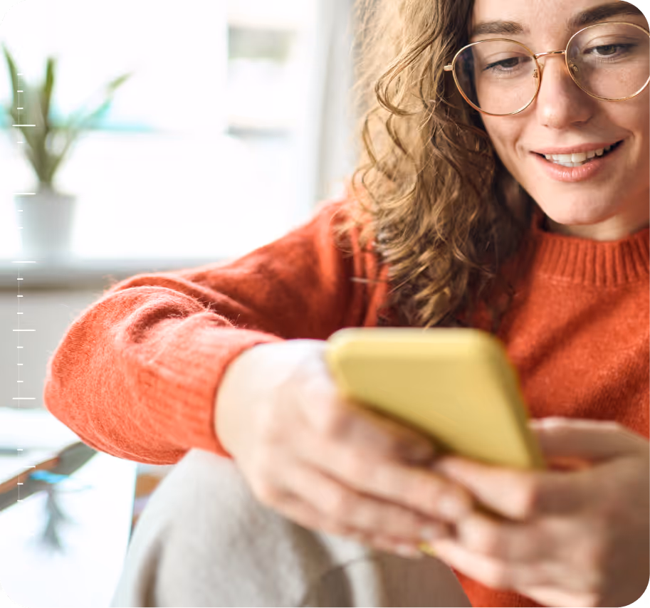 Smiling woman in glasses and orange sweater holding and looking at a yellow smartphone.