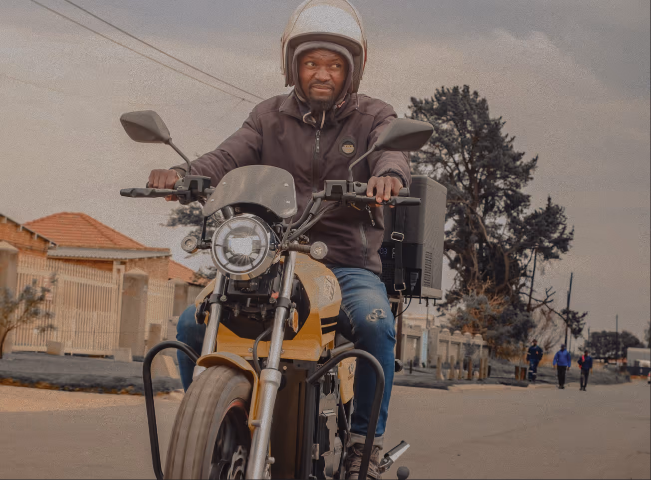 Man wearing helmet and dark jacket riding a yellow motorcycle on a suburban street with houses and trees.