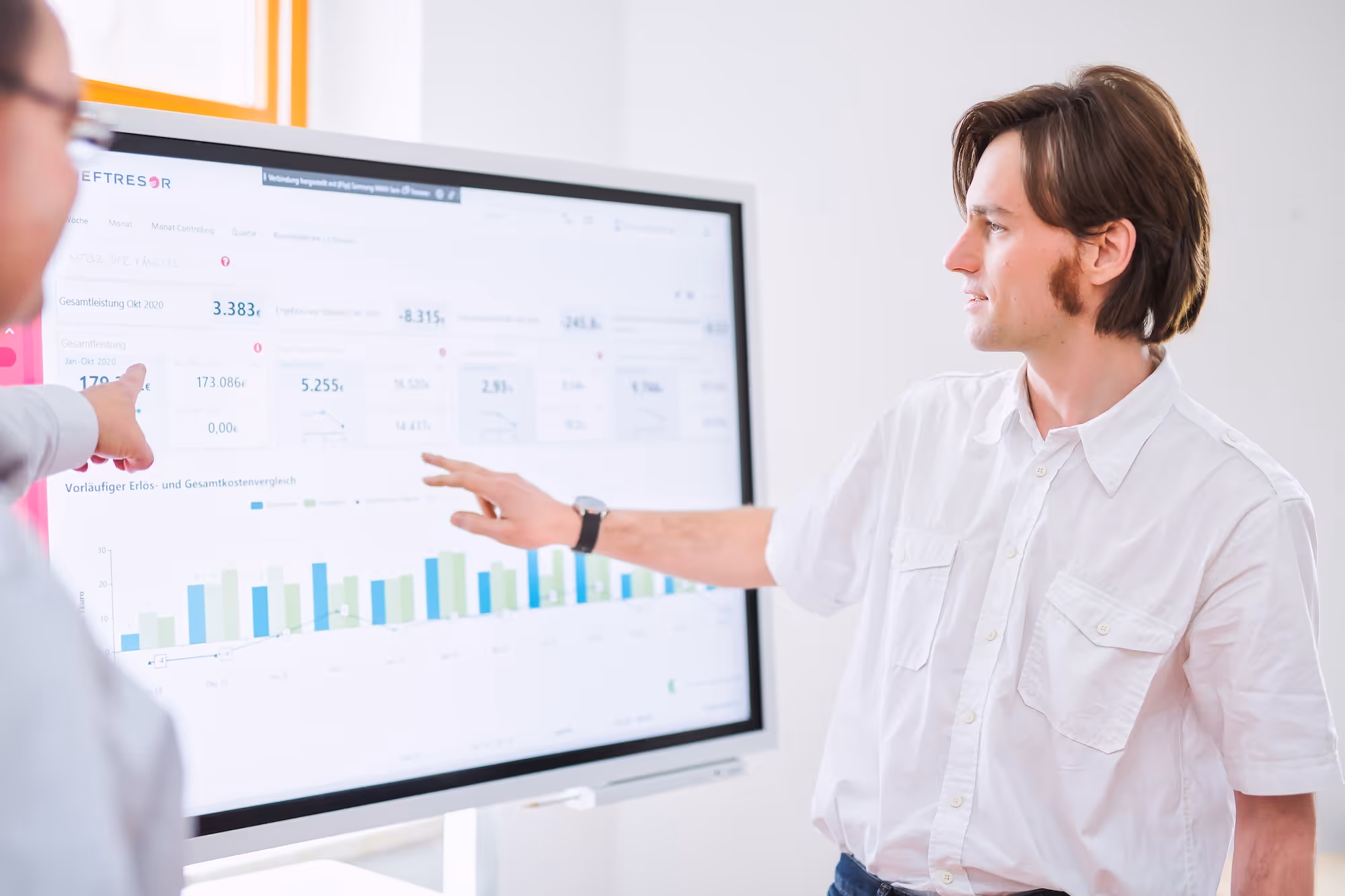 Zwei Männer diskutieren Finanzdaten und Diagramme auf einem großen Bildschirm im hellen Büro.