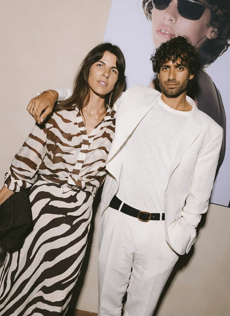 A man and a woman pose together indoors; the man wears a white blazer, white pants, a white shirt, and a black belt, with curly hair and a beard. The woman wears a long zebra-striped brown and white dress and holds a black clutch, with long brown hair. The man has his arm around the woman's shoulders. A large photo of a person wearing dark sunglasses is partially visible in the background.