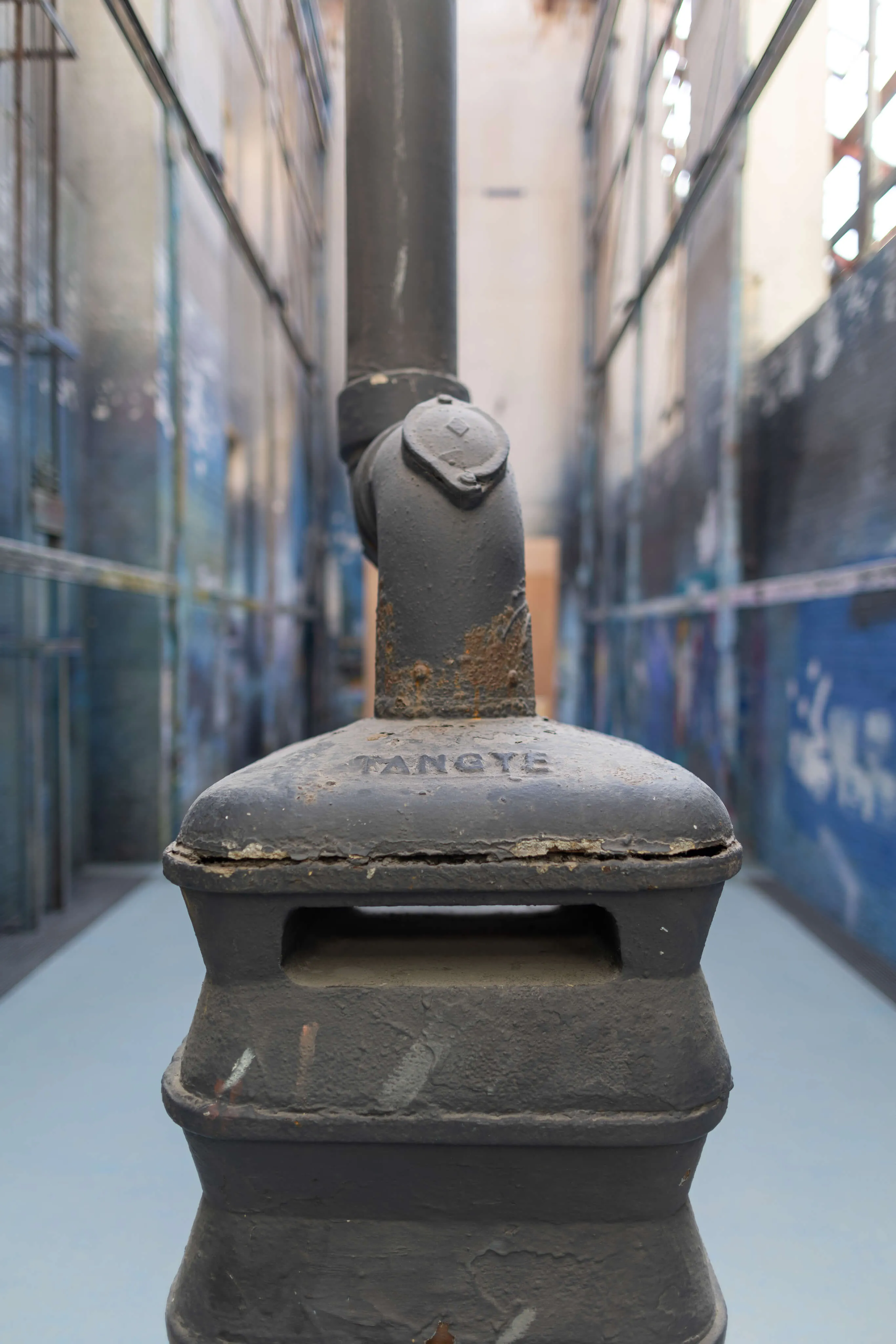 Close-up of an old, rusted black metal stove or heater pipe with the word 'YANGTE' embossed on the top, set inside an industrial space with blurred walls featuring peeling paint and metal framework.