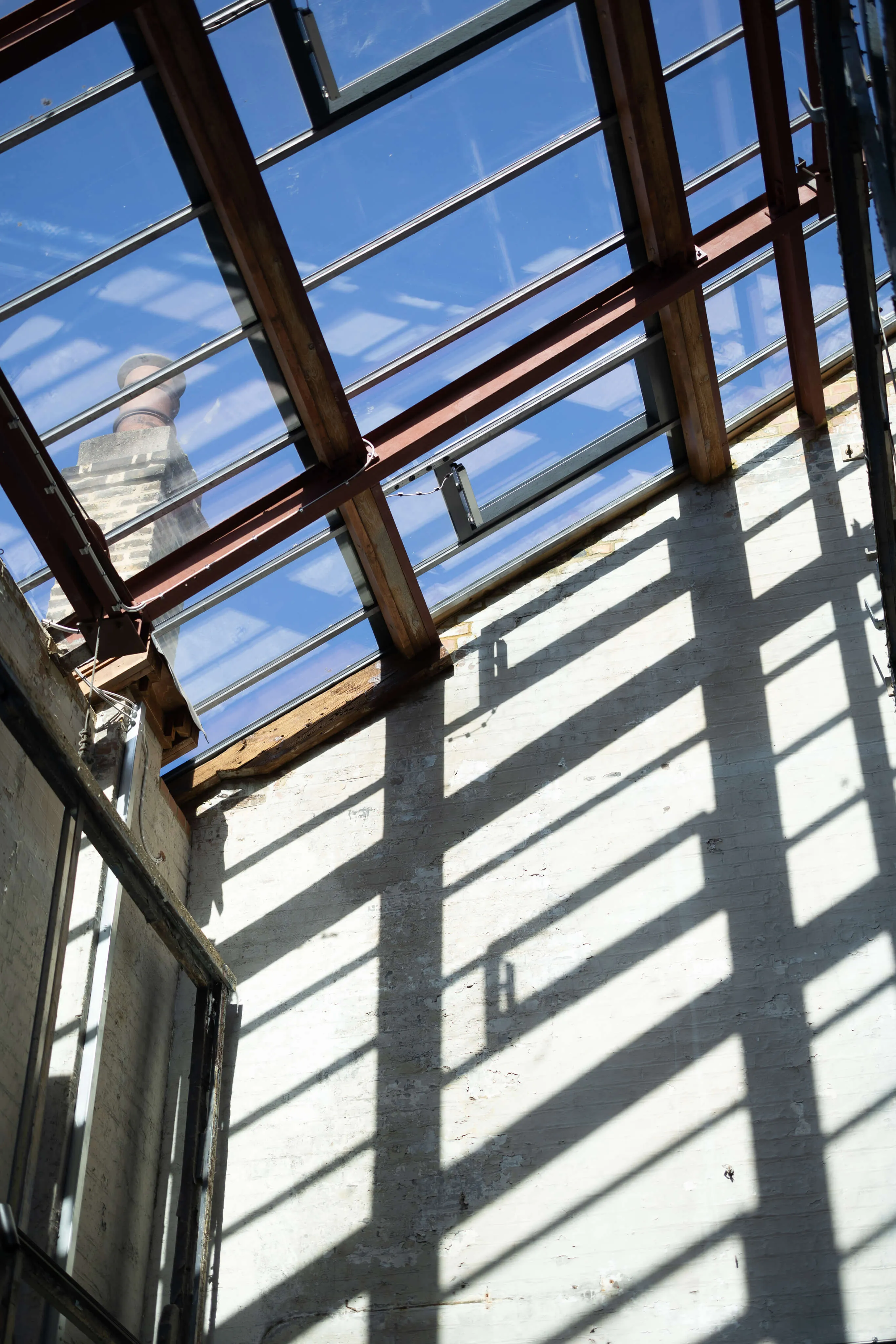 Interior view of a room with a glass ceiling supported by wooden beams casting grid-like shadows on a white brick wall under bright daylight.