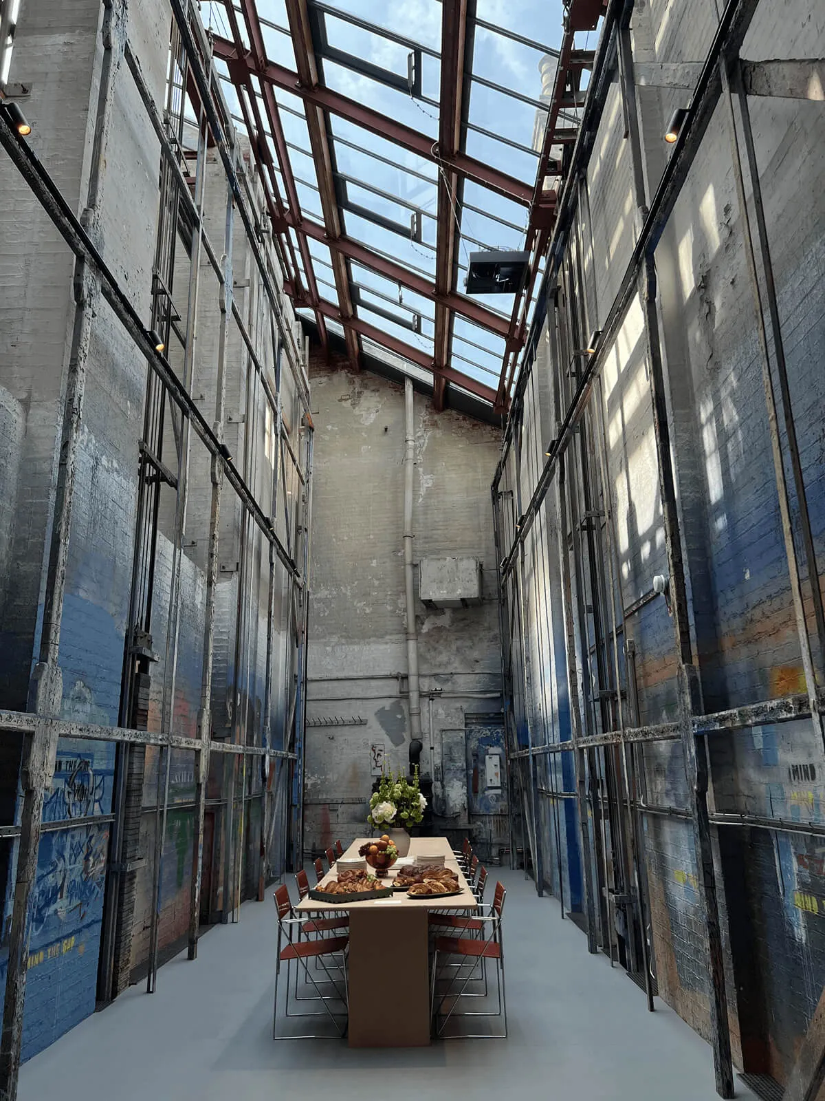 Long narrow industrial-style room with tall walls and a glass ceiling, featuring a wooden table in the center surrounded by red chairs, topped with pastries and a floral centerpiece.