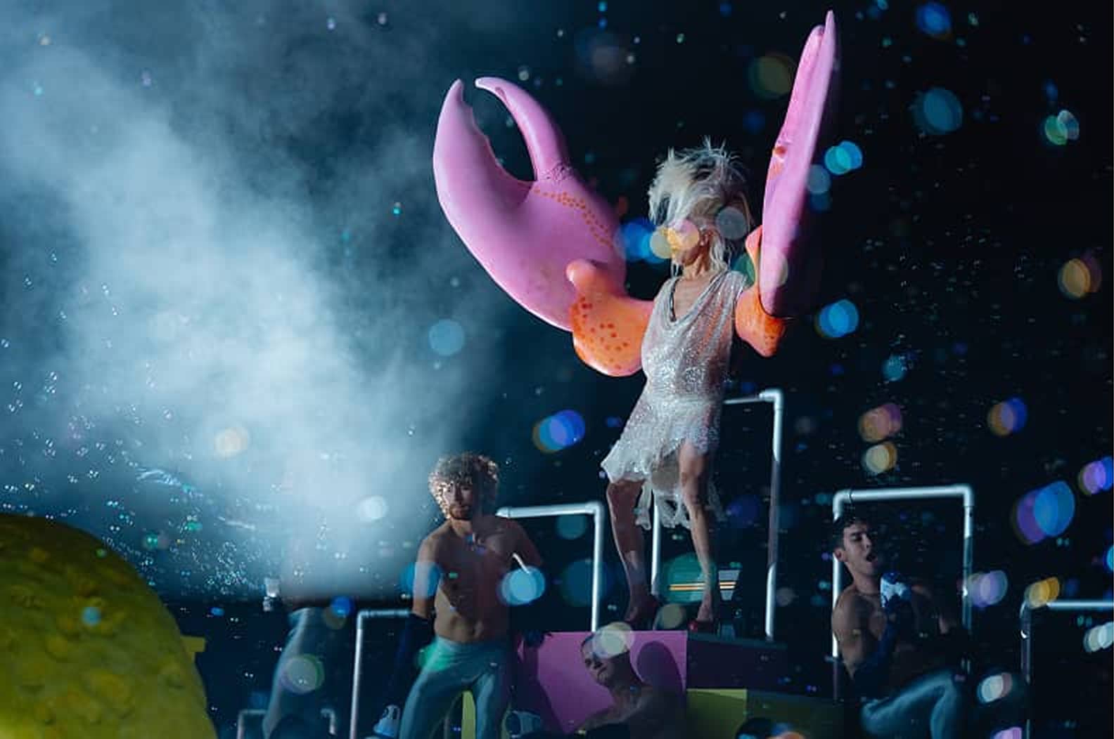A person wearing a sparkling dress and large pink crab claw gloves stands on a platform surrounded by three shirtless individuals under colorful stage lights and fog effects.