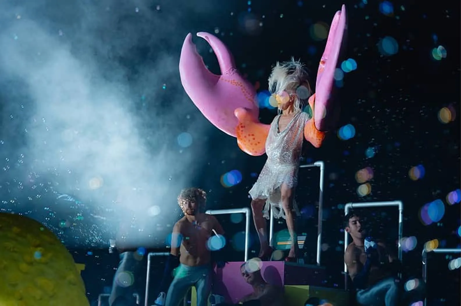 A person wearing a sparkling dress and large pink crab claw gloves stands on a platform surrounded by three shirtless individuals under colorful stage lights and fog effects.