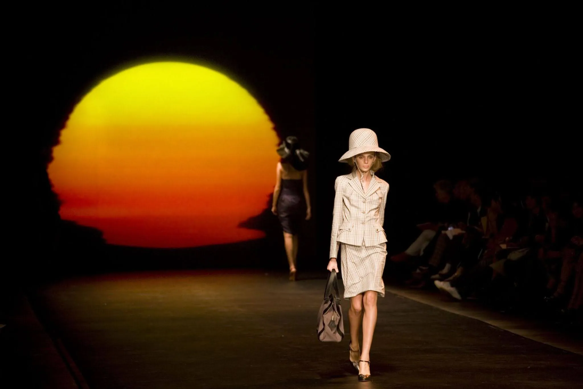 Fashion runway with two models; the one in front wears a beige plaid skirt suit, wide-brimmed hat, and carries a tote bag, walking towards the camera against a backdrop of a large yellow and orange sunset image.