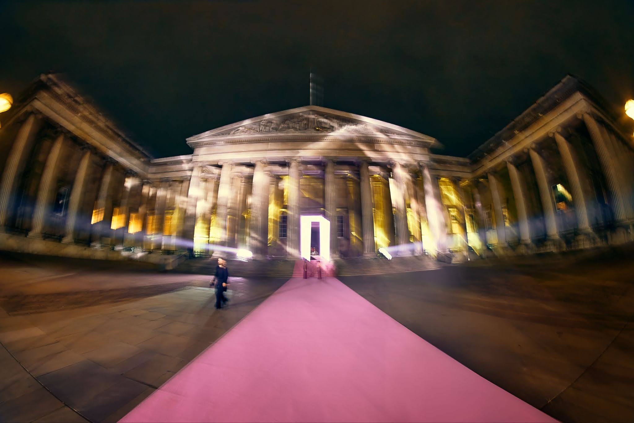 Night view of a large neoclassical building with tall columns lit up by yellow lights, a bright rectangular doorway, and a pink carpet leading to the entrance; a blurred figure walks on the left side near the carpet.