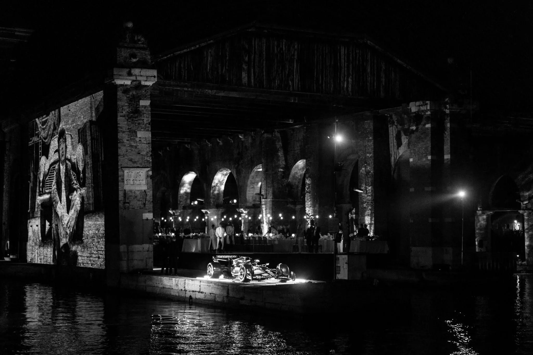 Black and white photo of a lit Formula 1 car displayed on a platform by water under a bridge with arched brick walls, people gathered in the background, and a large projected mural on the left wall.