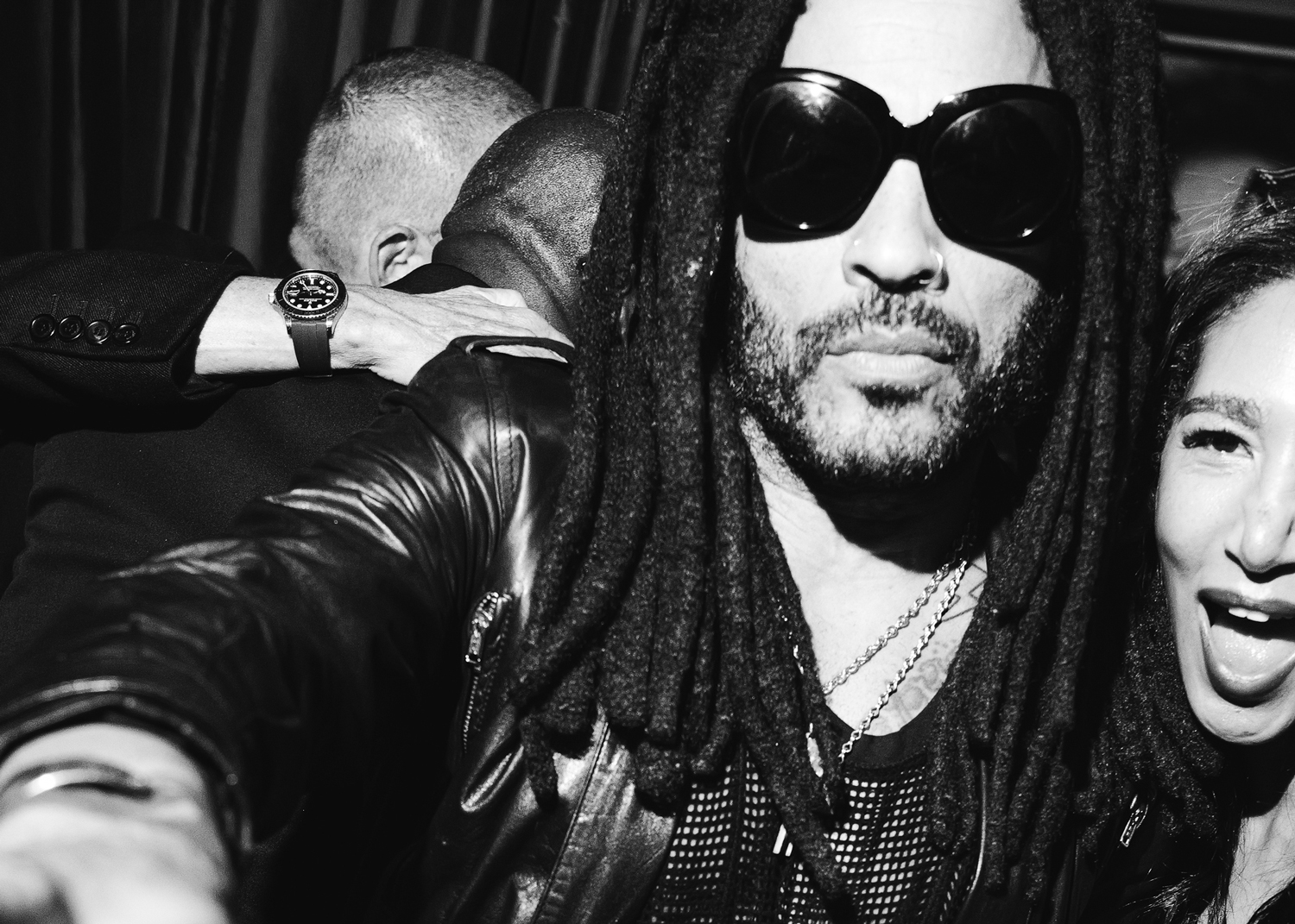 Black and white close-up photo of a man with dreadlocks wearing large sunglasses and a leather jacket, taking a selfie with a woman who is smiling and has her mouth open.