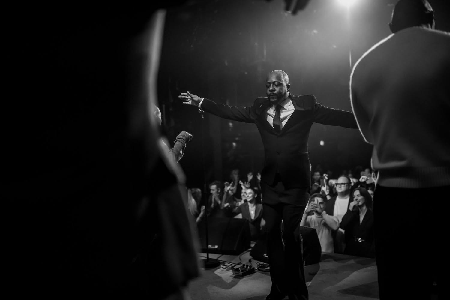 A man in a suit stands on stage with arms outstretched, eyes closed, in front of an applauding audience in a dimly lit venue.