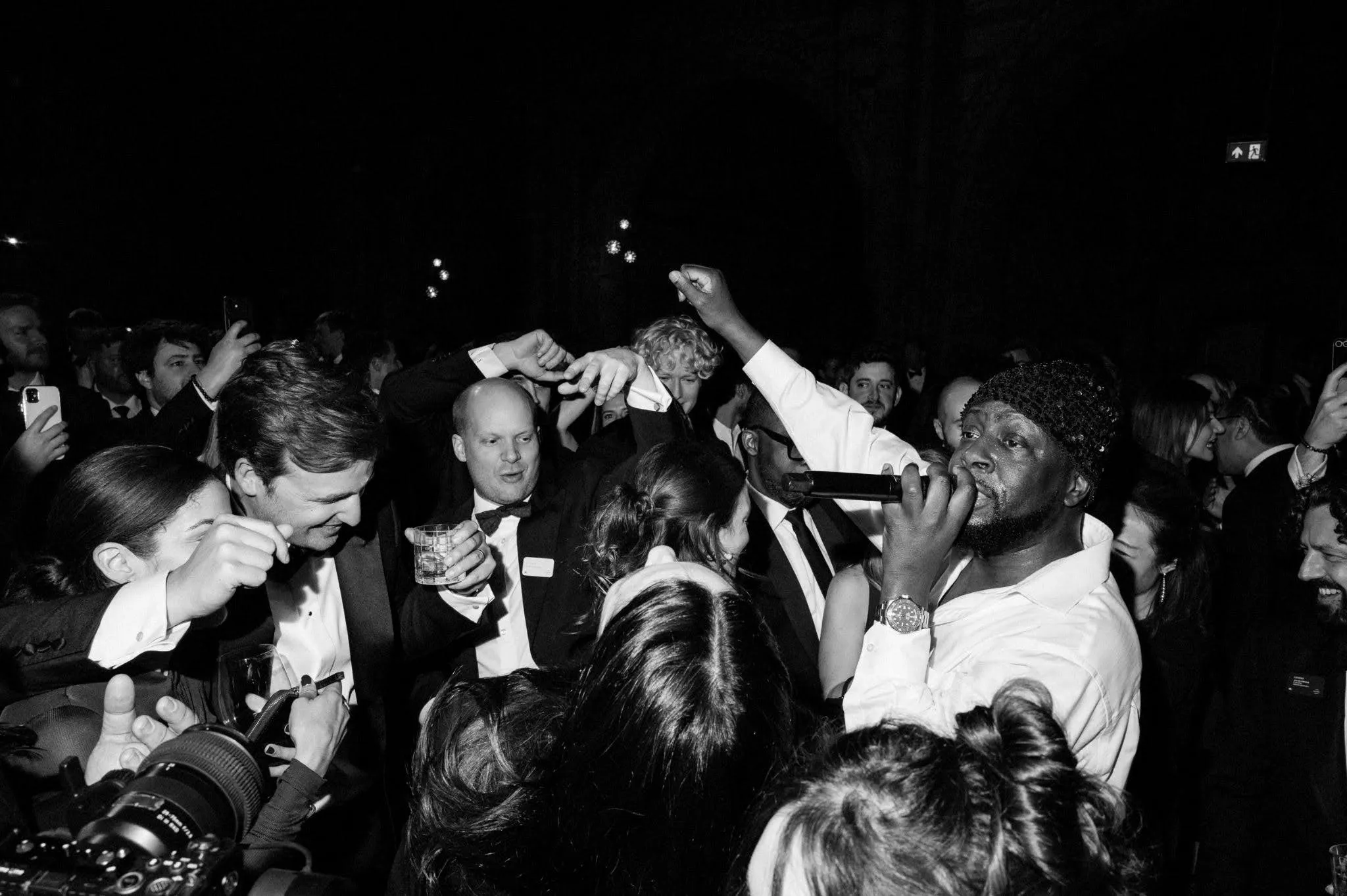 Black and white photo of a lively party scene with a man in a white shirt and knit cap singing or speaking into a microphone, surrounded by a crowd of people in formal attire dancing and enjoying themselves.