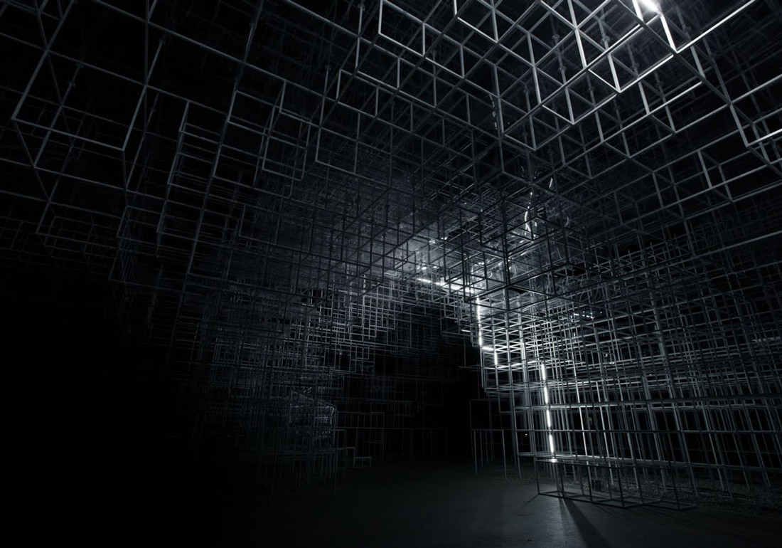 Dark room filled with an intricate three-dimensional grid structure made of interconnected metal or wire cubes, illuminated by a vertical strip of white light on the right side.