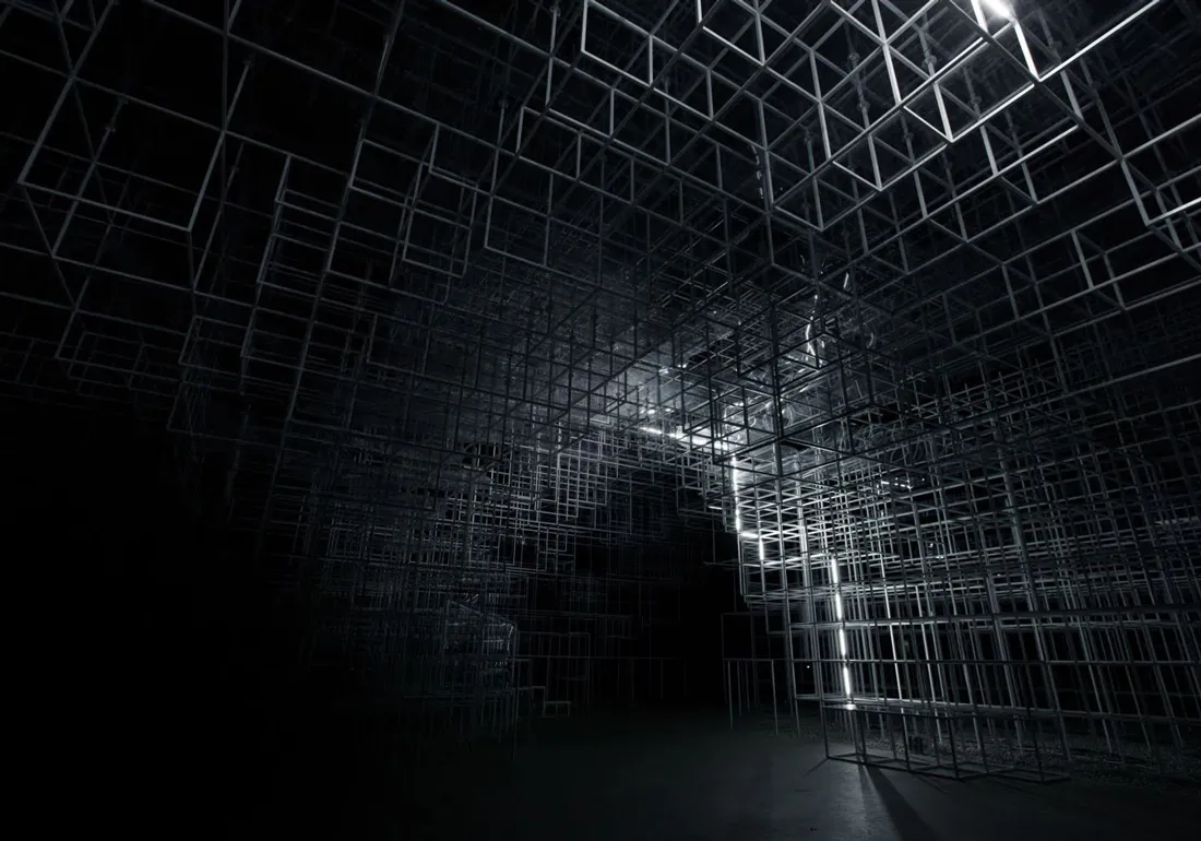 Dark room filled with an intricate three-dimensional grid structure made of interconnected metal or wire cubes, illuminated by a vertical strip of white light on the right side.