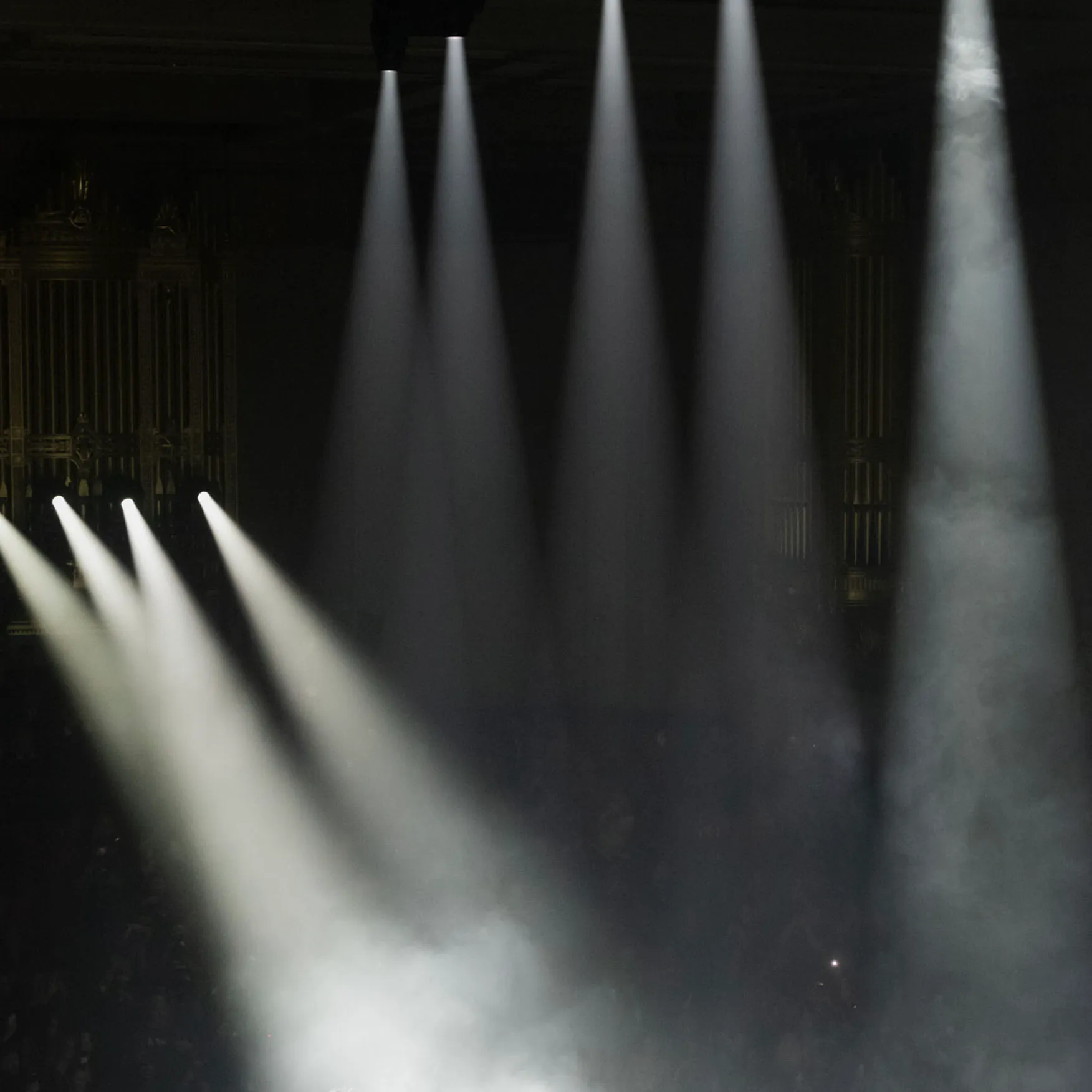 Multiple white spotlights beam downward through fog in a dark setting, suggesting a stage or concert environment.