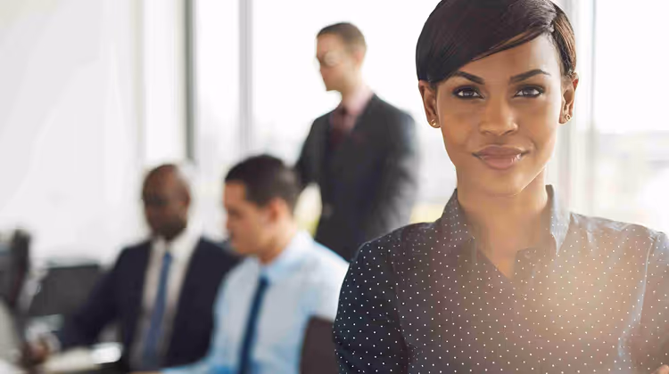 Confident businesswoman standing in the foreground with three coworkers blurred in the background in an office setting.