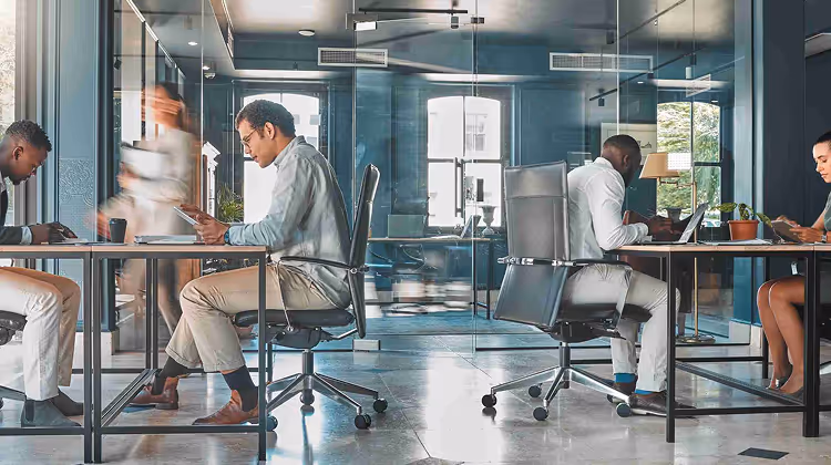 Four diverse professionals seated at desks in a modern office with glass walls, focused on their work while one person walks past.