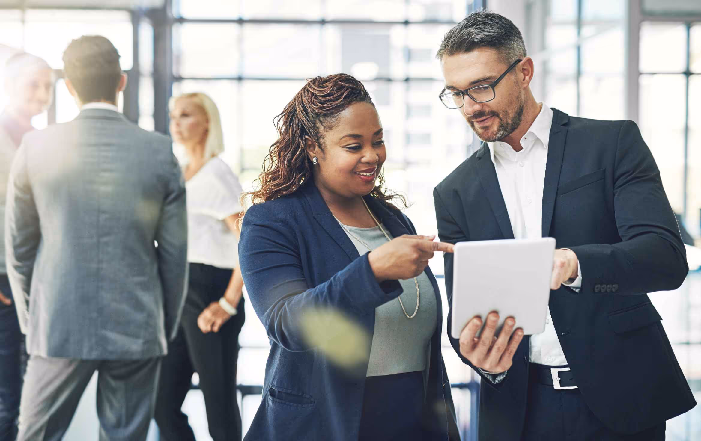 Two business professionals in suits discussing content on a tablet in a modern bright office.