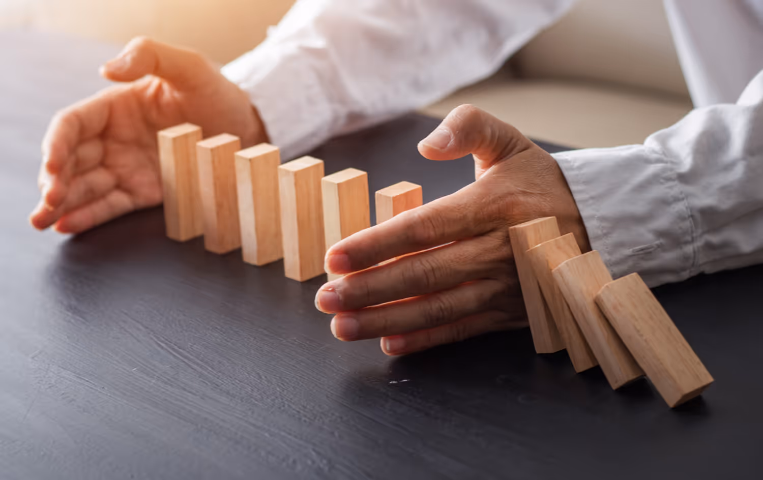 Hands preventing a row of wooden dominoes from falling on a dark surface.