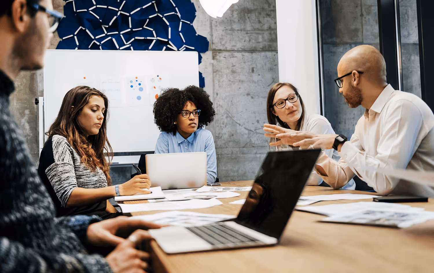 Group of diverse colleagues in a modern office having a discussion around a conference table with laptops and papers.