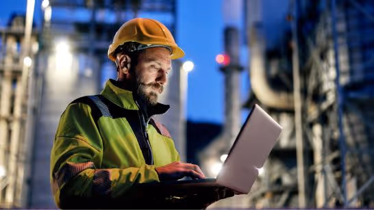 Engineer wearing a yellow hard hat and reflective jacket working on a laptop at an industrial site during dusk.