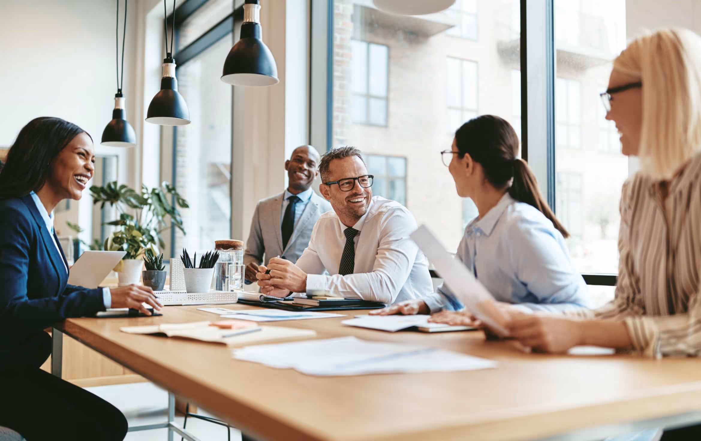 Five diverse professionals smiling and having a collaborative meeting around a wooden table in a modern office.