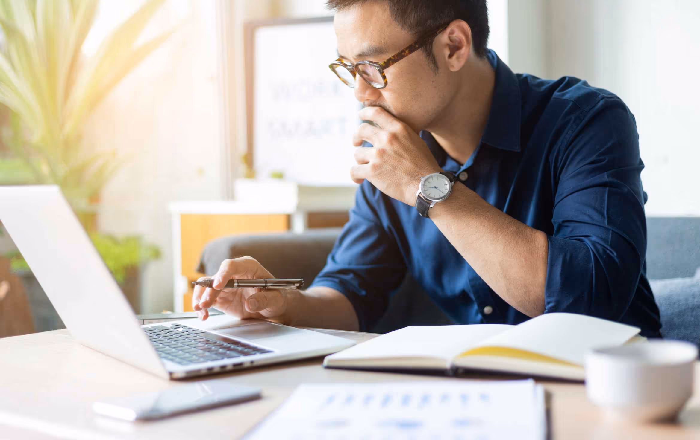 Man in glasses working on a laptop while holding a pen and drinking from a cup at a desk with an open notebook and documents.