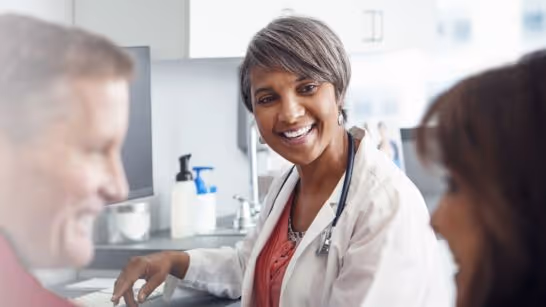 Smiling female doctor in white coat talking to two patients in a clinical setting.