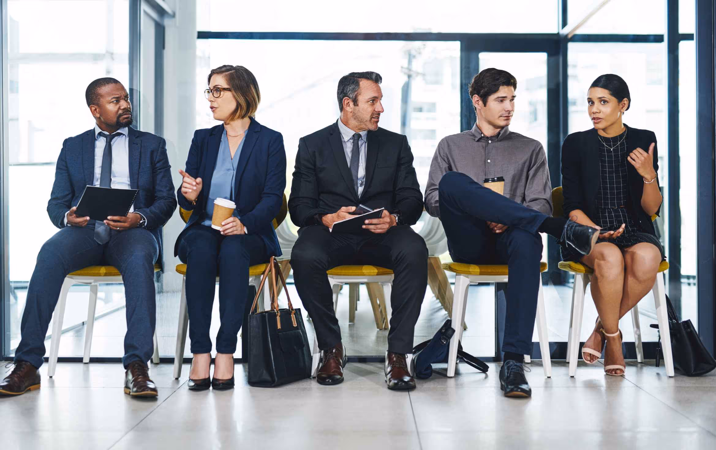 Five professional individuals sitting in a row in a bright office waiting area, engaged in conversation and holding notebooks or coffee cups.