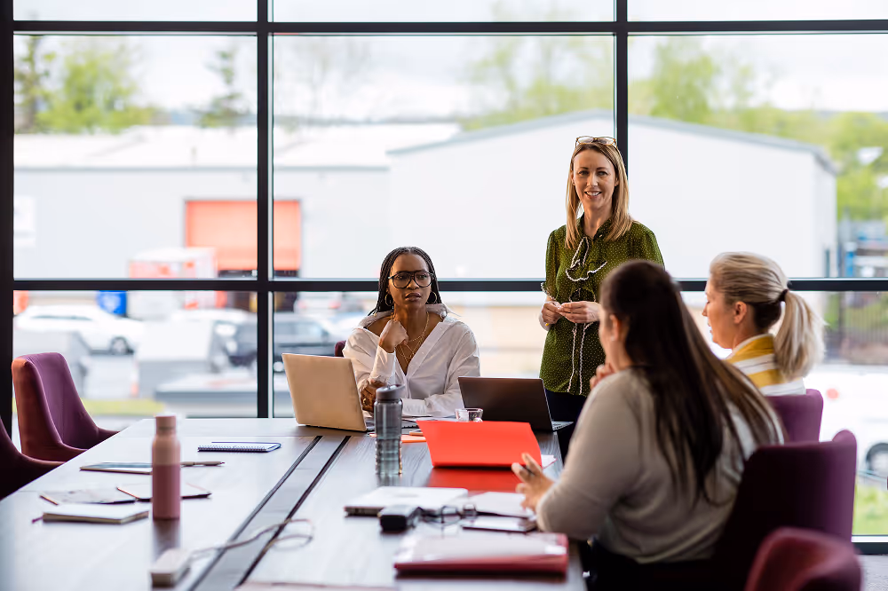 Four women in a meeting room with laptops and notebooks, discussing around a table by large windows.