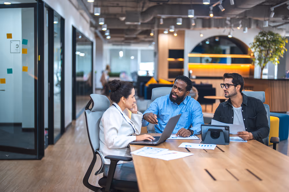 Three colleagues having a discussion around a table with laptop and documents in a modern office.